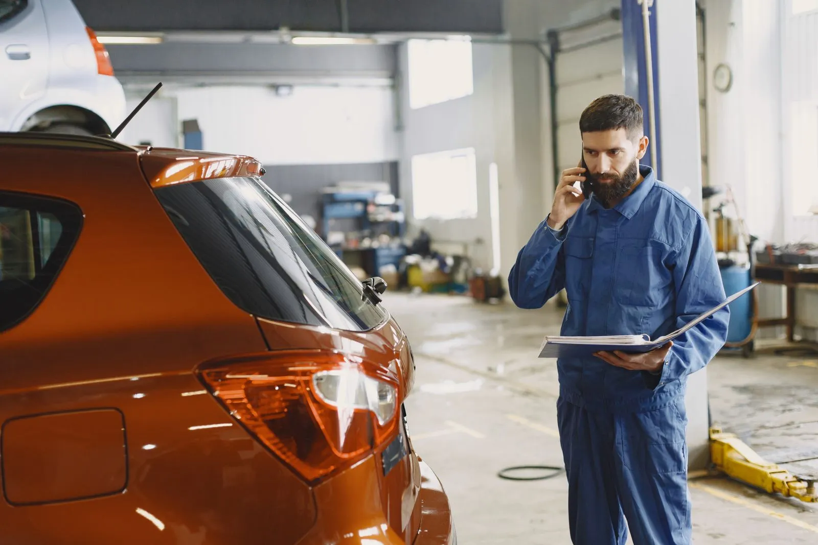 Meccanico dell'officina al telefono con la cartellina del preventivo in mano, accanto all'auto in lavorazione